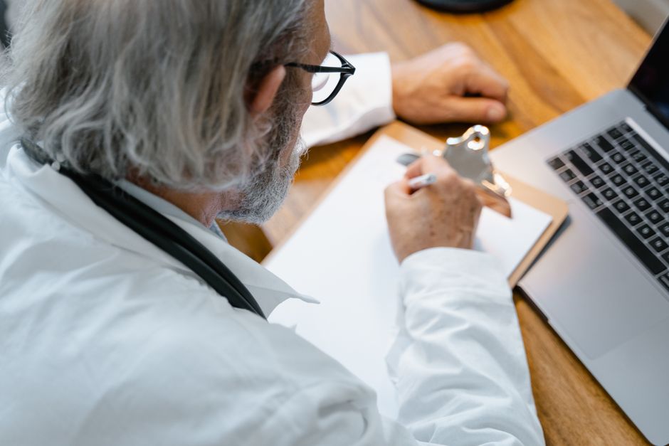 A homeopathic practitioner writing detailed case notes while listening attentively to a patient seated across a wooden desk in a calm, well-lit clinic room