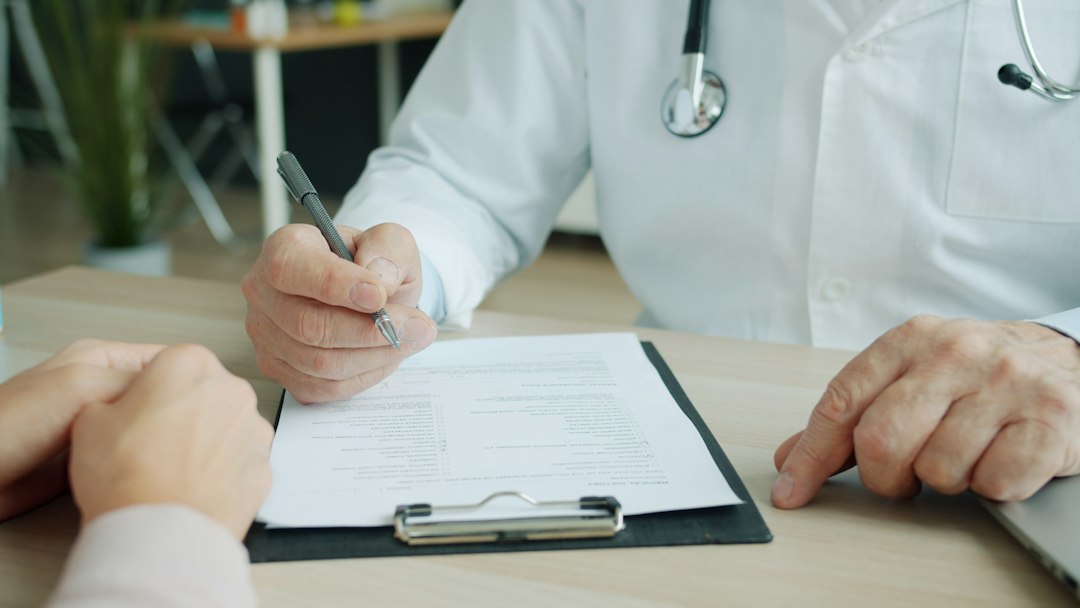 A homeopathic practitioner writing detailed notes while a patient describes digestive complaints during a case-taking session, focused and methodical